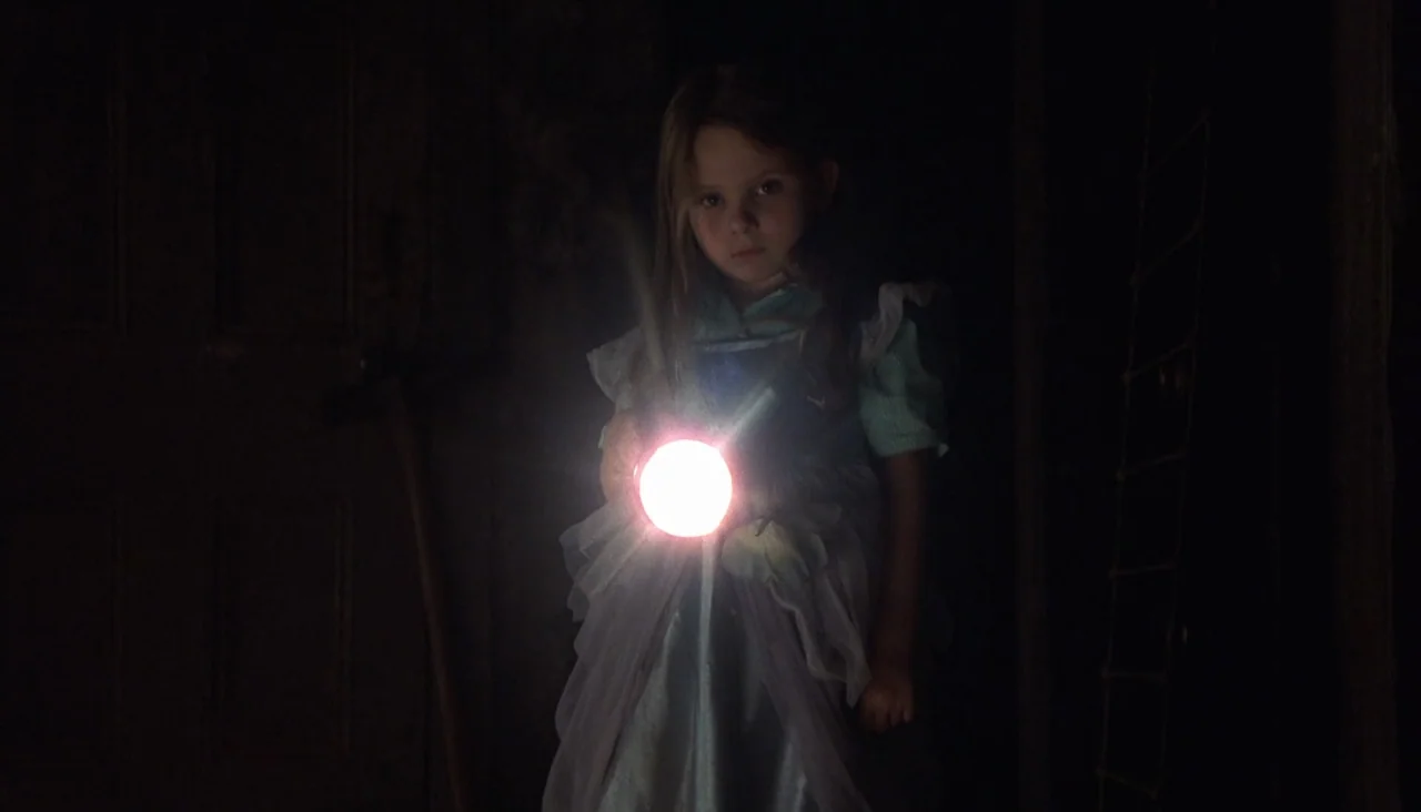 Signs (2002) movie still: medium — A young girl stands in a pitch-black, dimly perceived interior space with indistinct walls. She holds a lit…