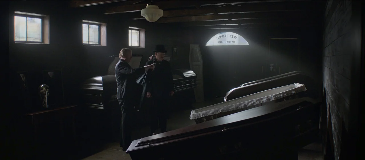 The Power of the Dog (2021) movie still: wide — A dimly lit room filled with rows of wooden caskets for a funeral home. Two men in formal black…