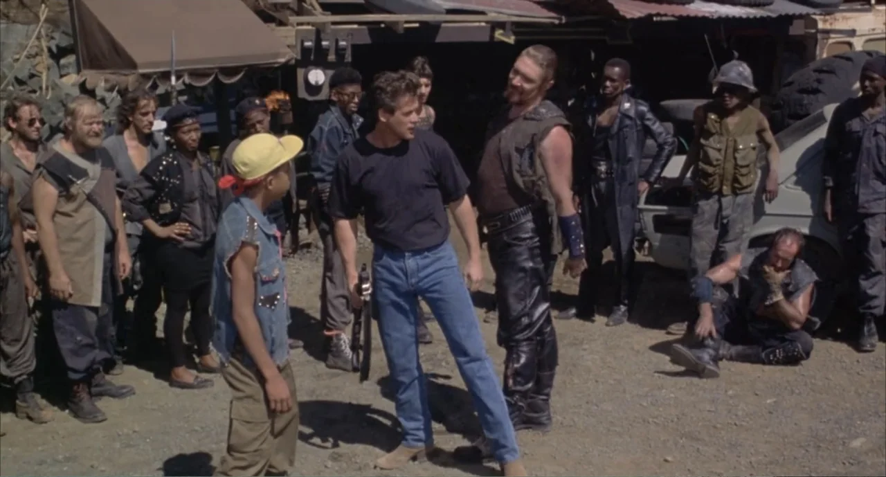 American Ninja 4: The Annihilation (1990) movie still: fullbody — A dusty outdoor scrapyard features piled tires and mechanical wreckage under…