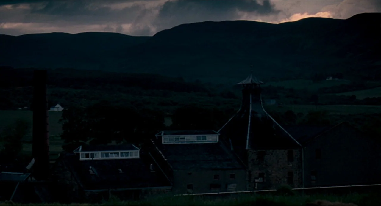 The Angel’s Share (2012) movie still: establishing — A stone distillery complex sits at the base of dark, rolling hills at dusk. A tall industrial…