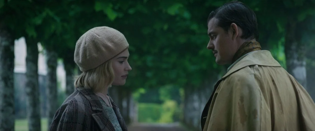 Rebecca movie still: medium — An outdoor pathway lined by tall trees creates a natural corridor of deep green foliage. A man and a woman stand…