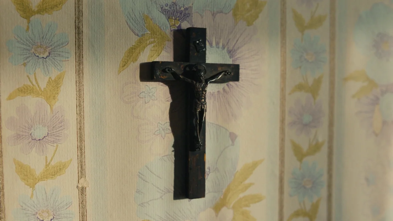 Lover’s Rock (2020) movie still: closeup — A dark wooden crucifix hangs on a wall with vintage floral patterned wallpaper. The crucifix casts a…