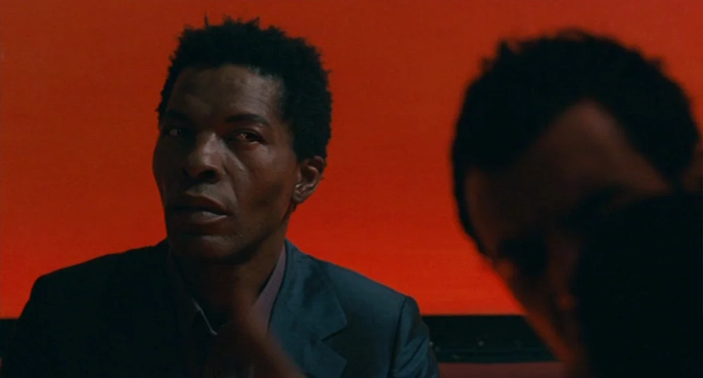 The Limits of Control (2009) movie still: medium — A man sits in front of a solid, bright red wall in a dark space. He wears a dark suit and looks…
