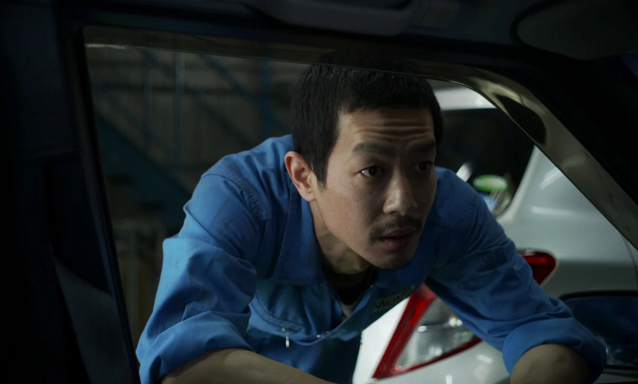 Like Someone In Love (2012) movie still: medium — An interior of an auto repair facility with a white vehicle partially visible behind. A male…