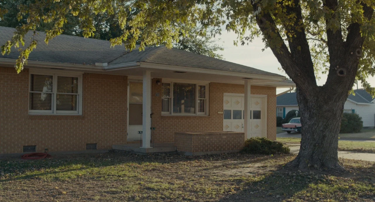 Wildlife (2018) movie still: establishing — A single-story tan brick suburban house sits under a large deciduous tree. A classic sedan is parked in…