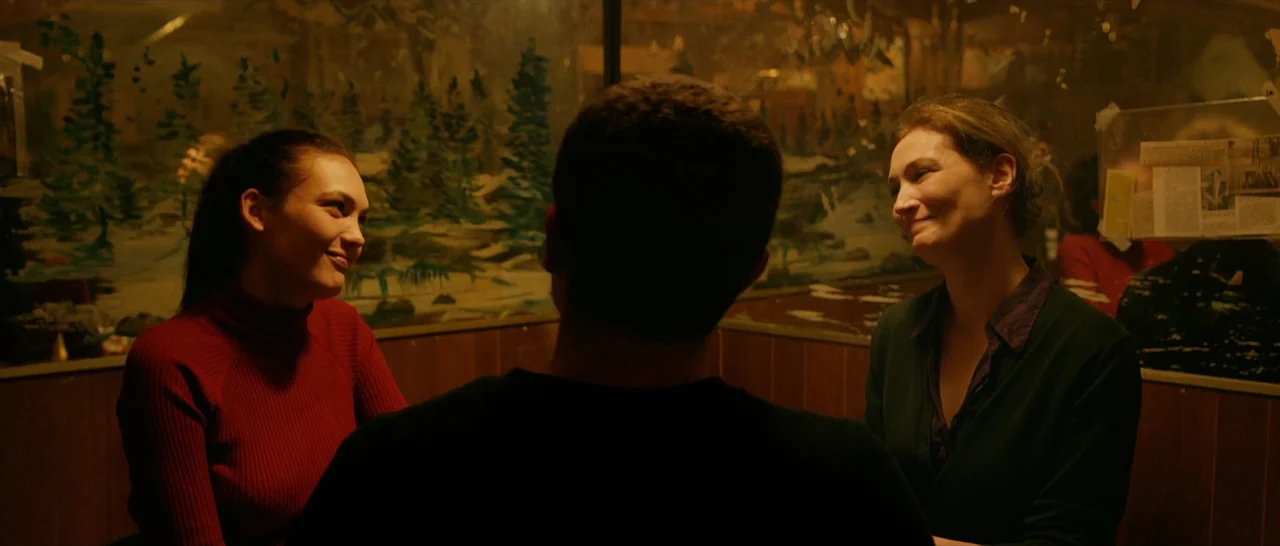 Love (2015) movie still: medium — A dimly lit diner booth features wood-paneled walls and a landscape painting in the background. Two women sit…