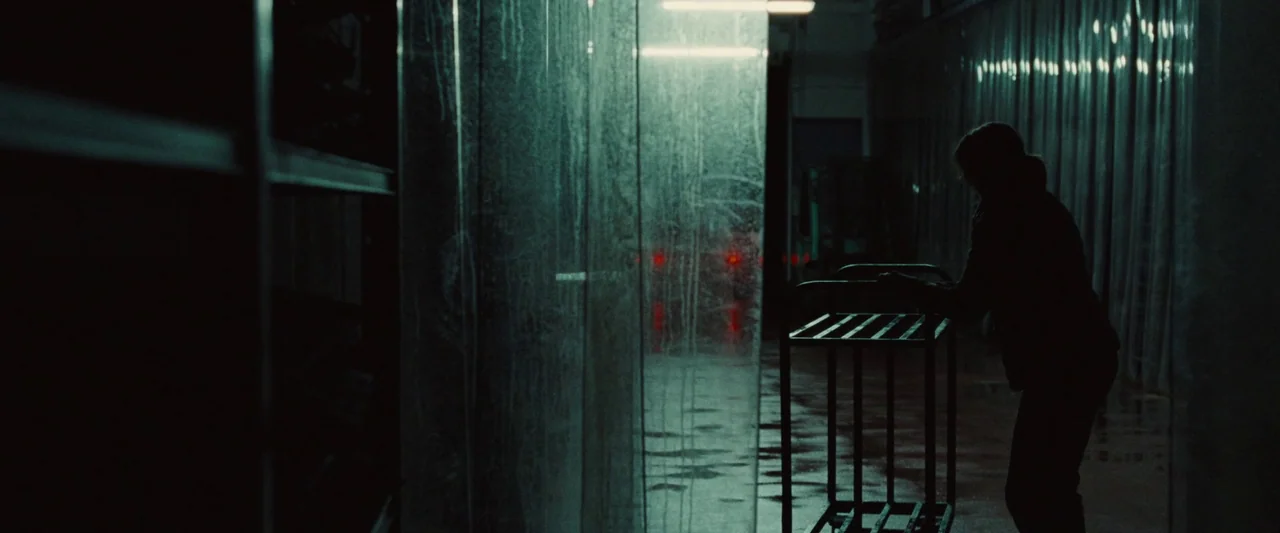 God’s Creatures (2022) movie still: medium — A dark warehouse aisle features cold, green-lit plastic curtains and wet, reflective flooring. A…