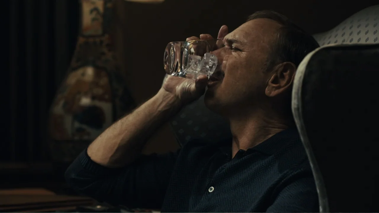 Paterno (2018) movie still: closeup — A dimly lit room with an ornate armchair and a patterned headrest. A man drinks water from a glass filled with…