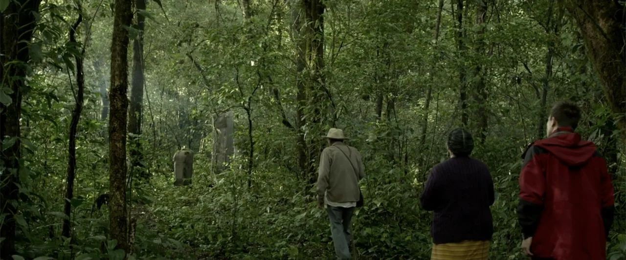 I Dream in Another Language (2017) movie still: wide — A dense subtropical forest with heavy foliage and thin, vertical tree trunks. Three people…