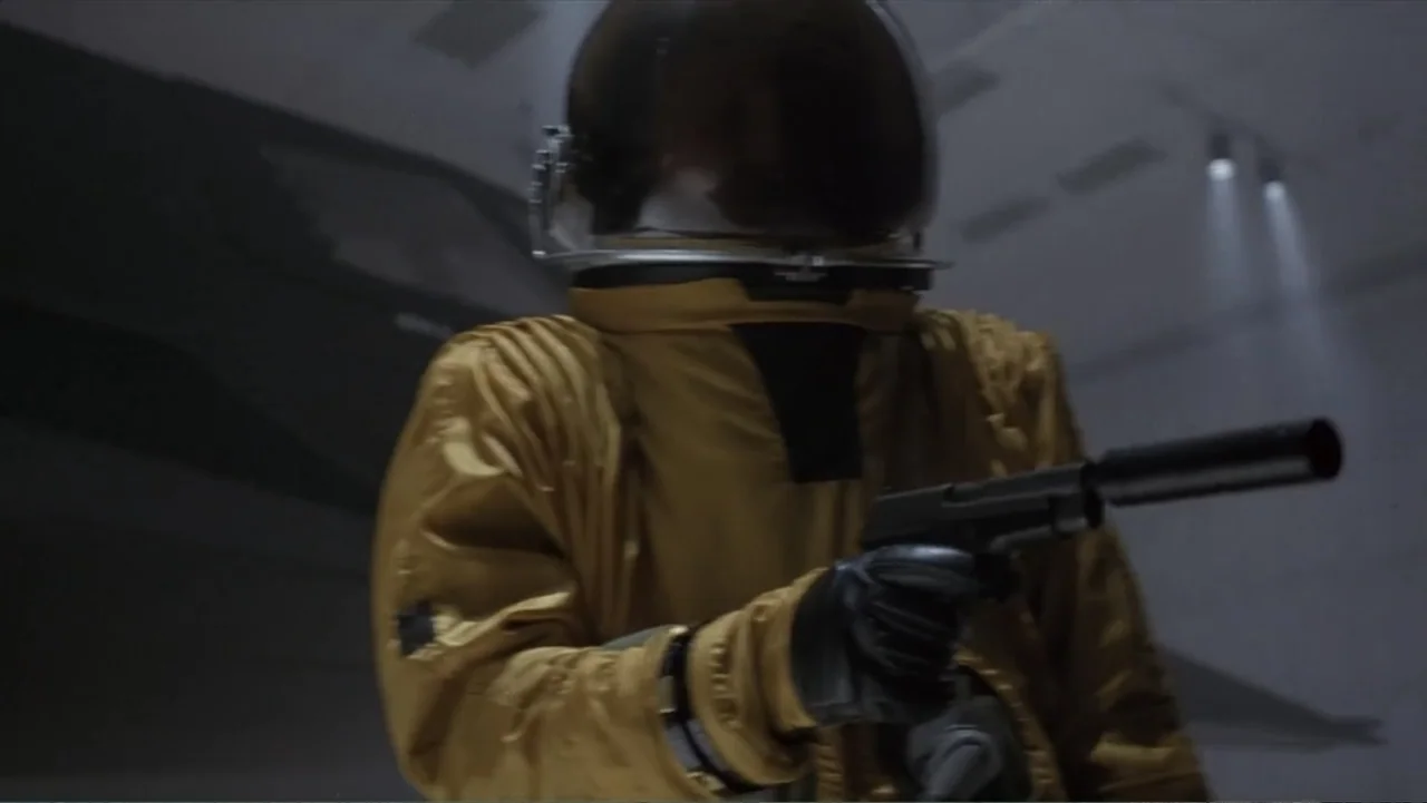 Crisis Point (2012) movie still: medium — A dimly lit industrial room features concrete surfaces and muted geometric shadows. A person in a golden…