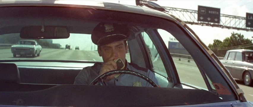 Brewster McCloud (1970) movie still: medium — The interior of a moving 1970s patrol car on a sunny highway. A police officer drives while talking…