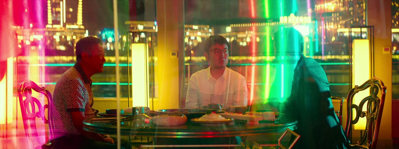 Under The Light (2023) movie still: medium — A glass-enclosed dining area overlooks a city skyline at night lit by bright neon. Three men sit around…