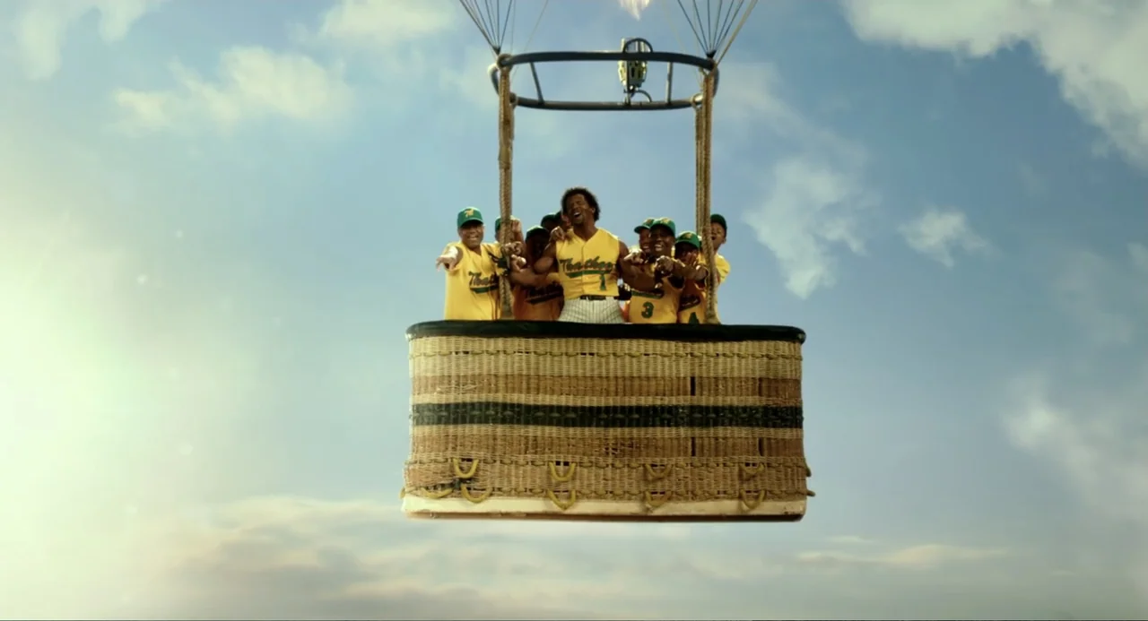 Blended (2014) movie still: wide — A woven hot air balloon basket floats against a bright blue sky with clouds. A group of men in yellow baseball…