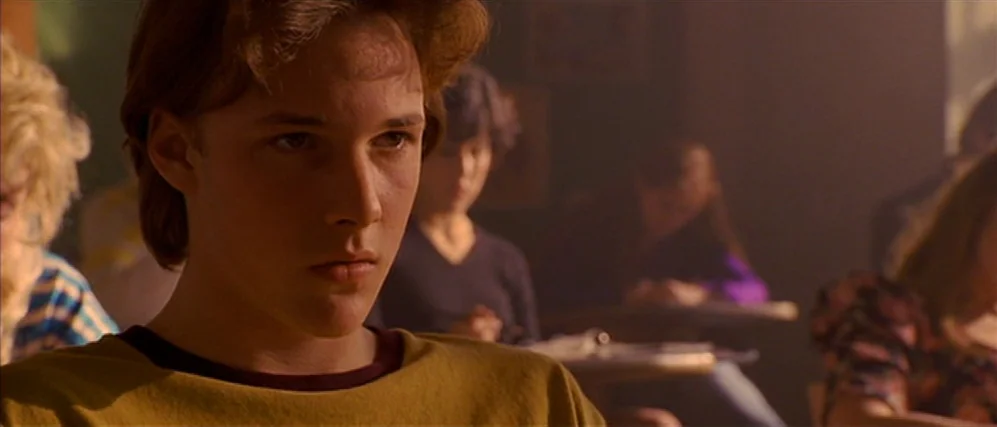 Apt Pupil (1998) movie still: closeup — A high school classroom filled with students sitting at desks in the background. A teenage boy with wavy…