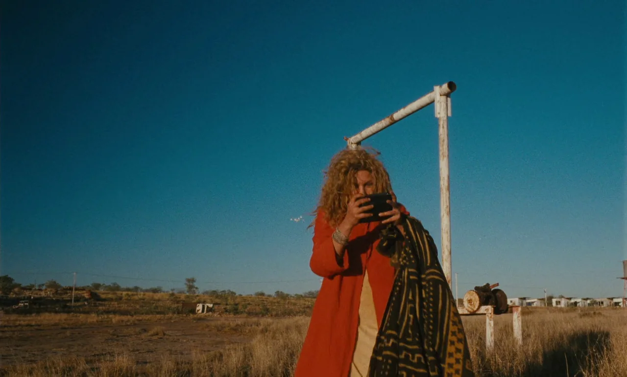 Until The End Of The World (1991) movie still: medium — A dry, grassy field under a cloudless blue sky contains a white metal frame. A woman with…