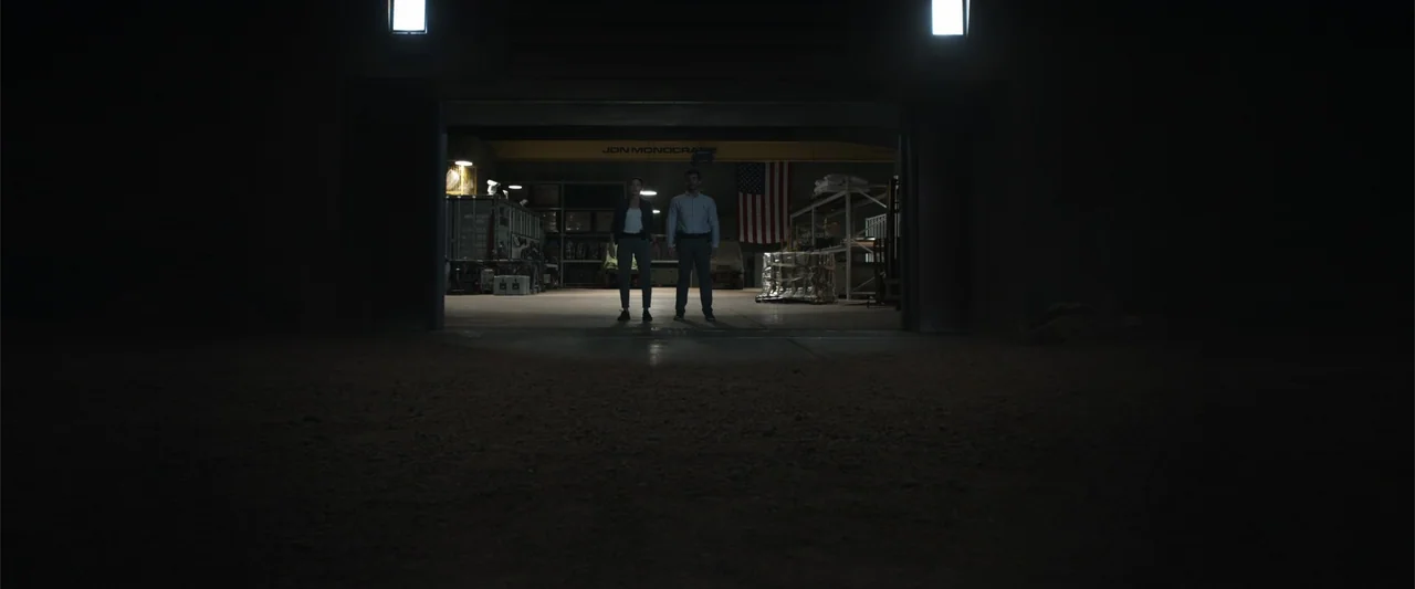 Black Site (2022) movie still: wide — A dark warehouse entrance is seen from a gravel exterior at night. A man and a woman stand inside the lit…