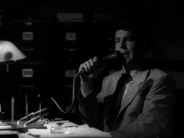 Double Indemnity (1944) movie still: medium — A dark office features metal filing cabinets in the background behind a desk. A man in a suit speaks…