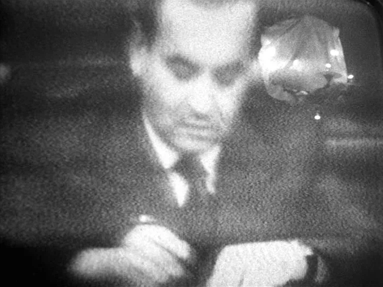 Family Nest (1979) movie still: medium — An interior space with a dimly lit desk environment. A man in a suit writes while a lamp shines from the…