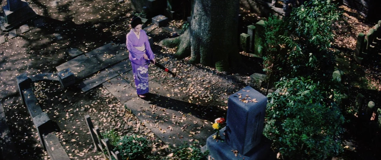 Lady Snowblood (1973) movie still: wide — A quiet, leaf-strewn cemetery with ancient stone markers under a large tree. A woman in a purple kimono…