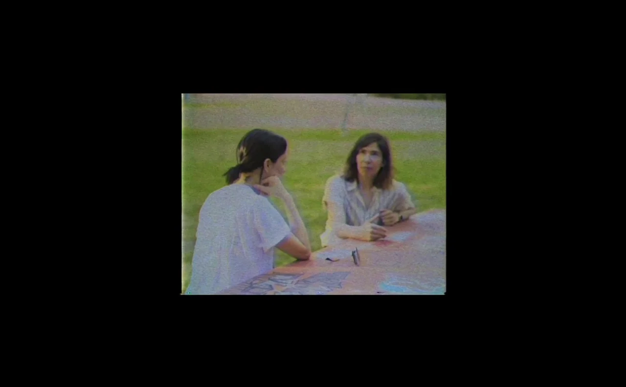The Nowhere Inn (2020) movie still: medium — Two women sit at a long wooden table in a grassy outdoor area. They converse while sitting face-to-face…