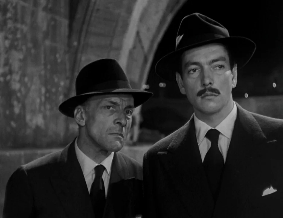 Roman Holiday (1953) movie still: medium — Two men stand near a dark stone archway at night. They look ahead with serious expressions while wearing…