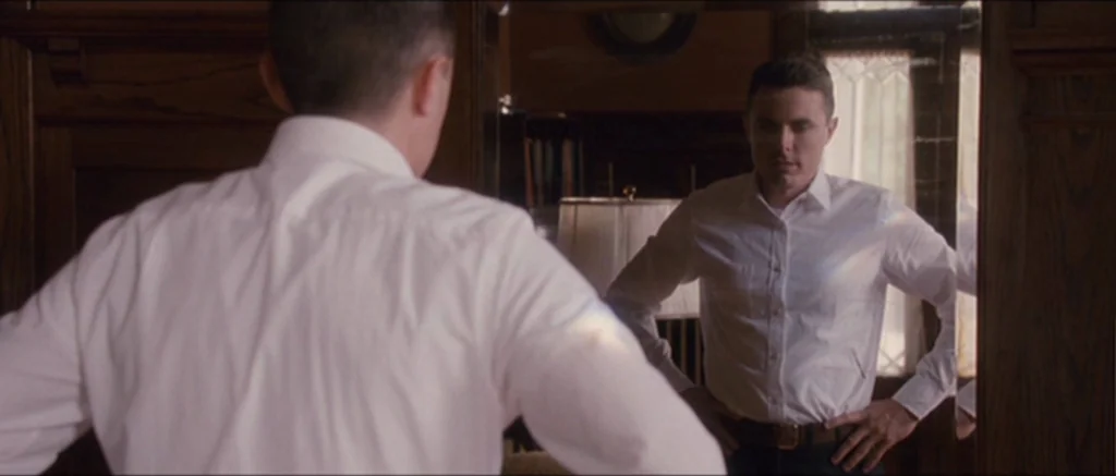 The Killer Inside Me (2010) movie still: medium — A man stands in a wood-paneled hallway facing a large mirror mounted on the wall. He observes his…