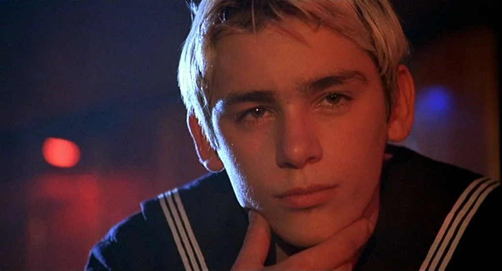 Deux (2002) movie still: closeup — A dimly lit room with blue and red neon lighting sources. A teenage male in a sailor outfit rests his chin on his…