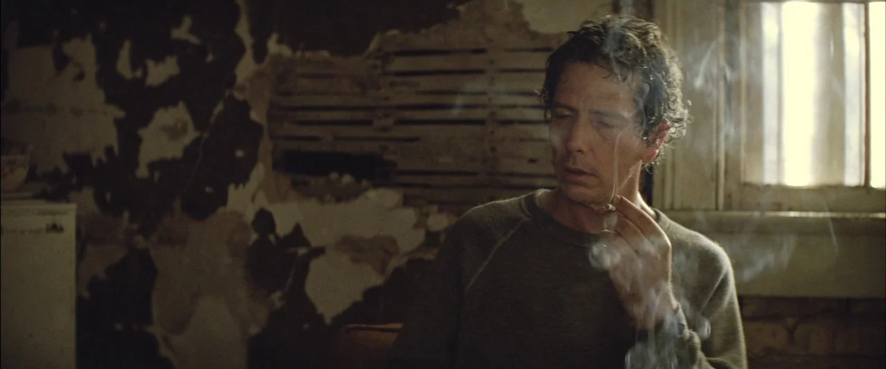 Killing Them Softly (2012) movie still: medium — A room with exposed wood lath behind peeling plaster walls near a window. A man holds a lit…