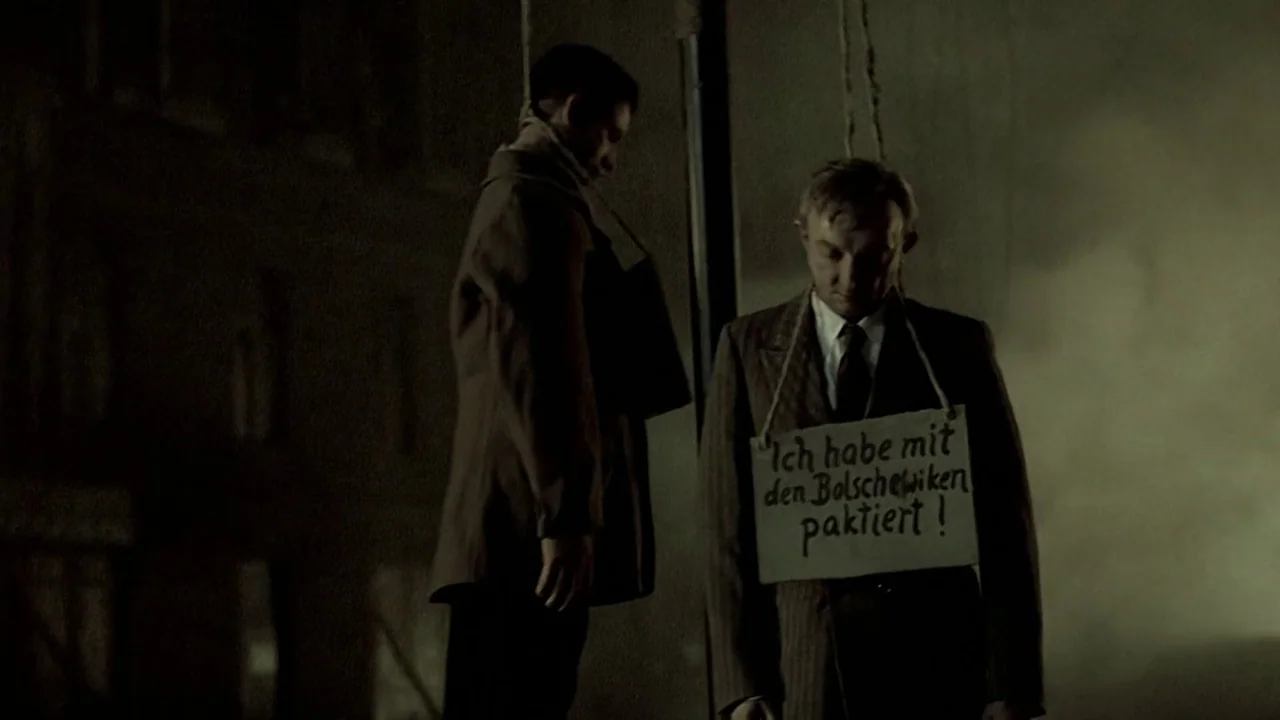Downfall (2004) movie still: medium — A dark, foggy urban street at night with two men hanging from ropes. The man in the foreground wears a sign…