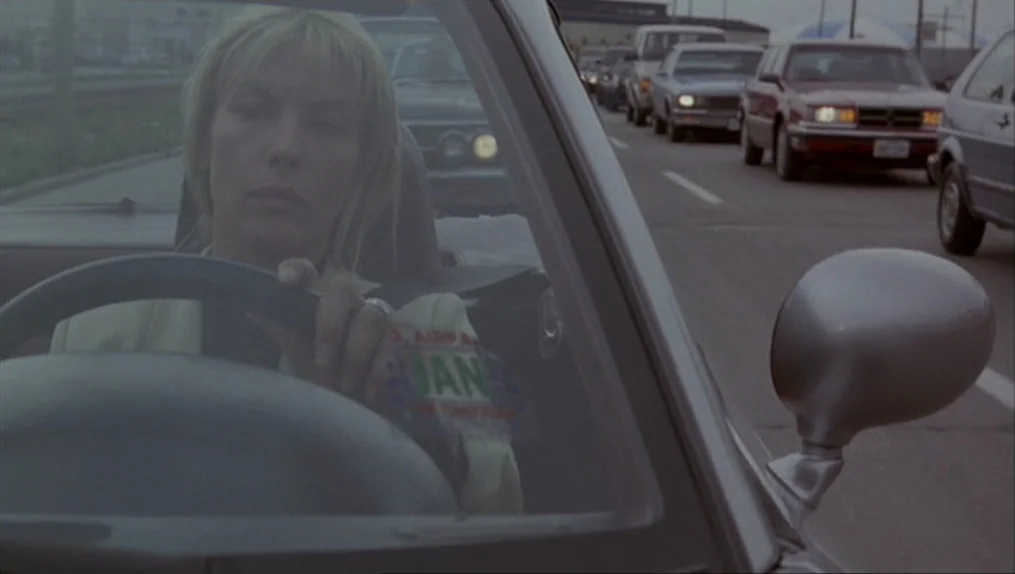 Crash (1996) movie still: medium — A woman drives her car through a busy multi-lane highway in overcast weather. Many other vehicles are visible in…