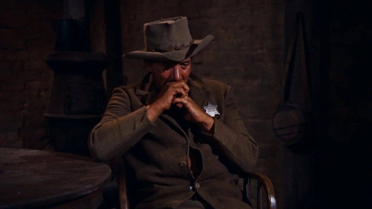 Rio Bravo (1959) movie still: medium — A dimly lit, rustic interior with visible brick walls and an iron stove behind. A man wearing a sheriff's…