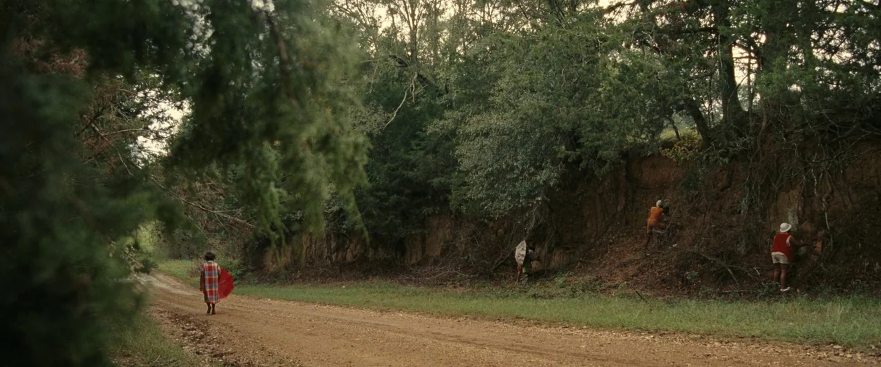 All Dirt Roads Taste of Salt (2023) movie still: wide — A rural dirt road passes a steep earthen bank topped by dense trees. Three figures occupy…
