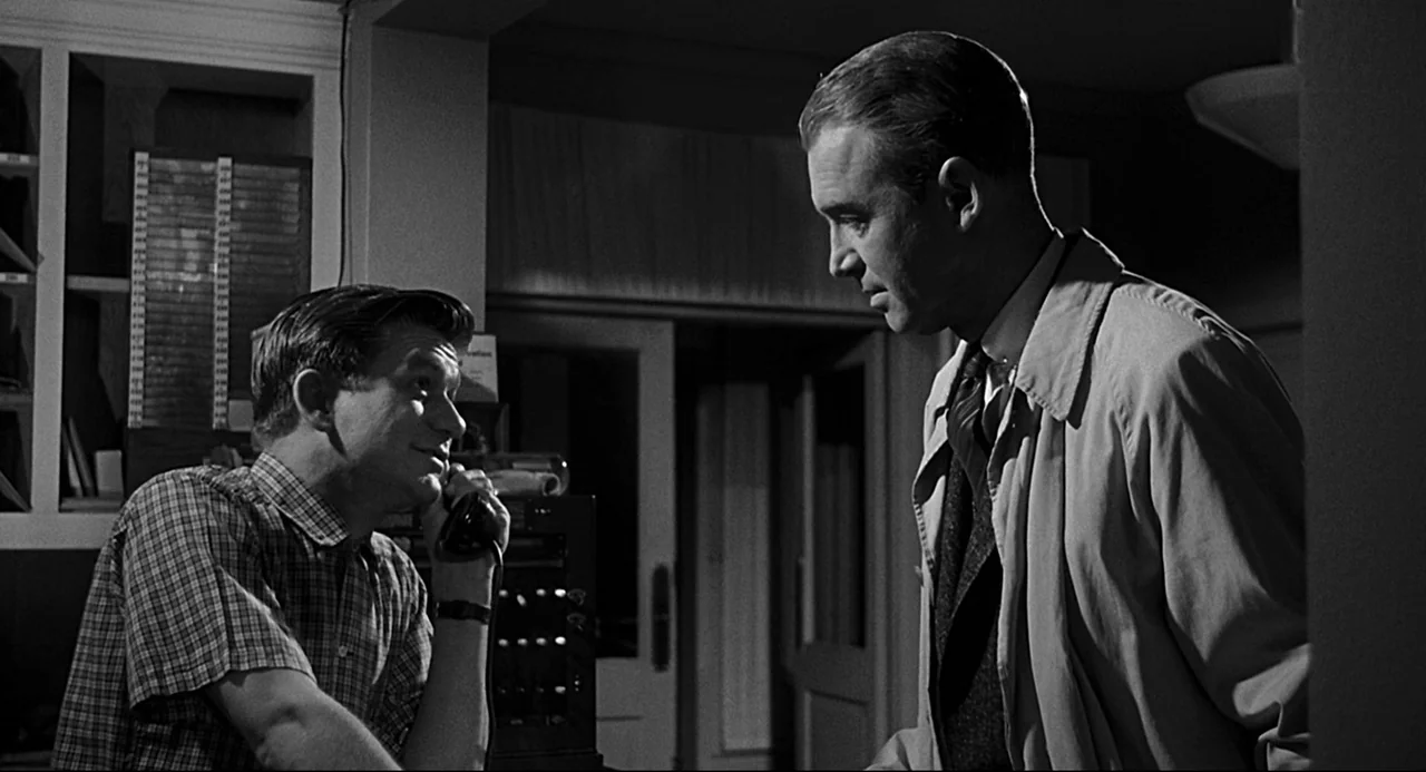 Anatomy of a Murder (1959) movie still: medium — A dimly lit room with filing shelves and telephone equipment in the background. One man speaks into…