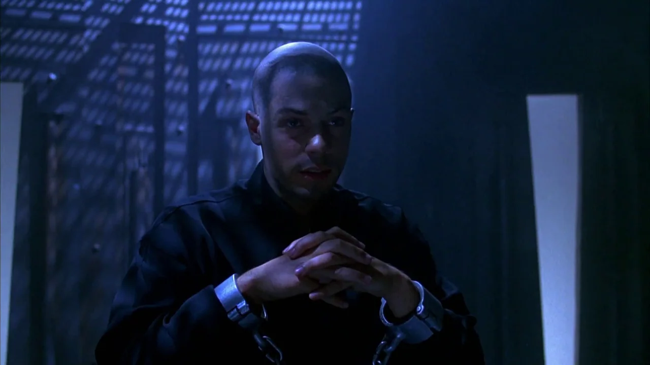 Hellraiser: Bloodline (1996) movie still: medium — A stark room features a metallic grate backdrop casting a grid pattern shadow. A man with a…