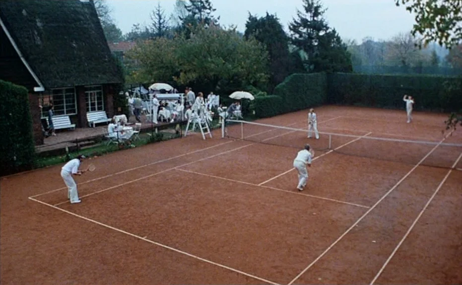 Soldier of Orange (1977) movie still: wide — A group of people participate in a social tennis match on clay. The court sits adjacent to a…