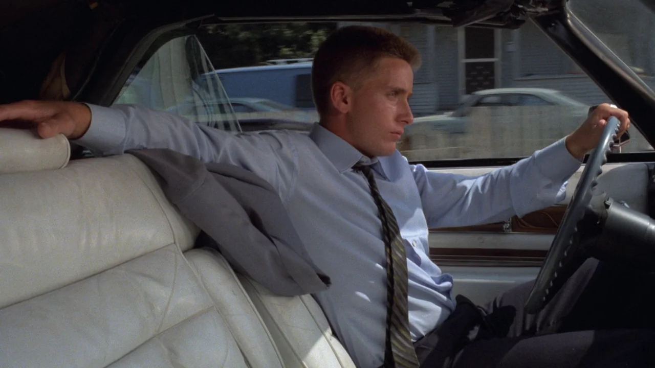 Repo Man (1984) movie still: medium — The interior of a vintage car features white leather bucket seats and a steering wheel. A man in a blue dress…