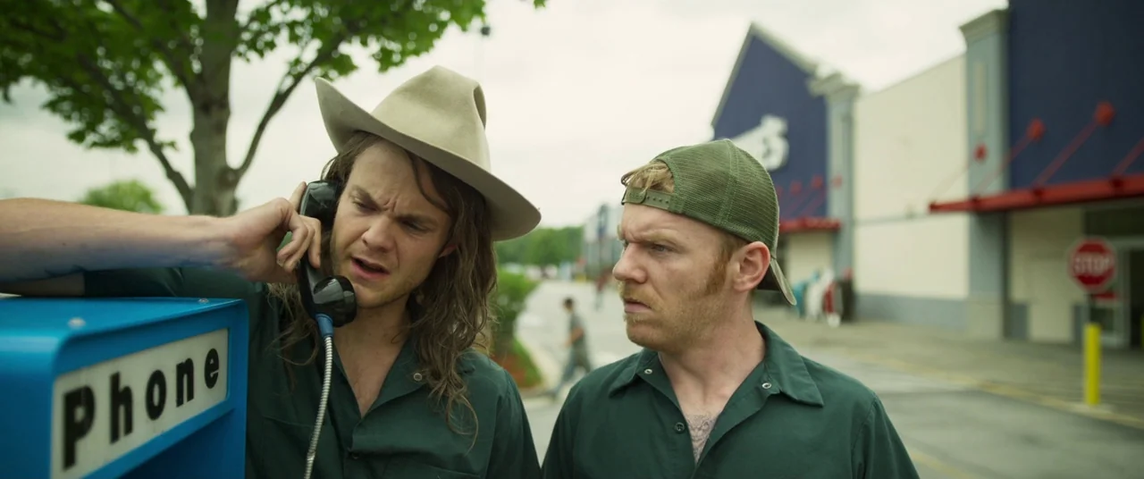 Logan Lucky (2017) movie still: medium — A man in a cowboy hat uses an outdoor payphone in a commercial parking lot. His associate stands next to…