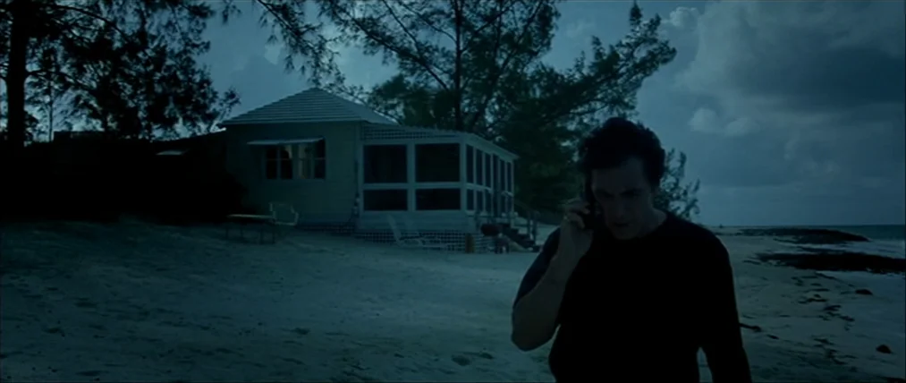 The Insider (1999) movie still: medium — A sandy beach at night with a small house in the background. A man walks forward along the shoreline while…