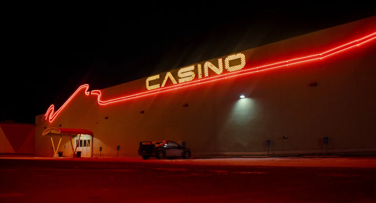 My Animal (2023) movie still: wide — A low-angle view of a beige concrete casino exterior wall at night. A dark coupe is parked in the foreground…