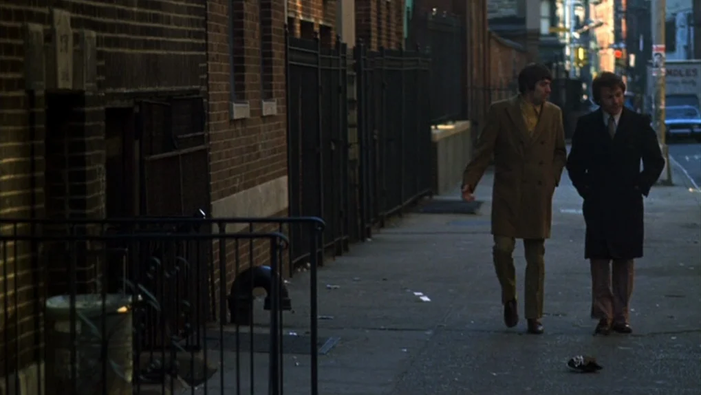 Mean Streets (1973) movie still: fullbody — A city sidewalk beside a brick building with iron railings and a metal trash can. Two men dressed in…