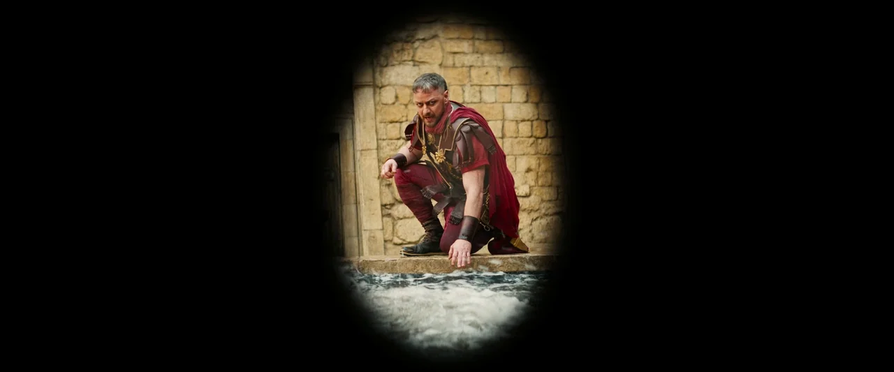 The Book of Clarence (2023) movie still: medium — A man in ancient Roman military attire crouches before a stone-bordered water feature. He reaches…