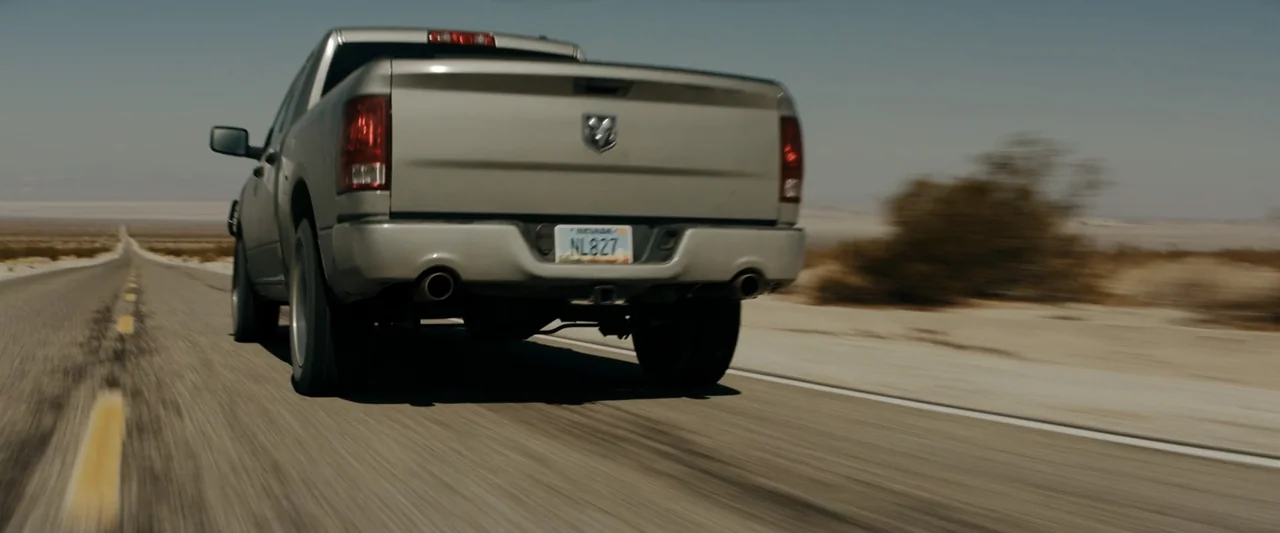 Encounter (2018) movie still: medium — A silver pickup truck travels down a long, asphalt road through a flat desert. The vehicle occupies the…