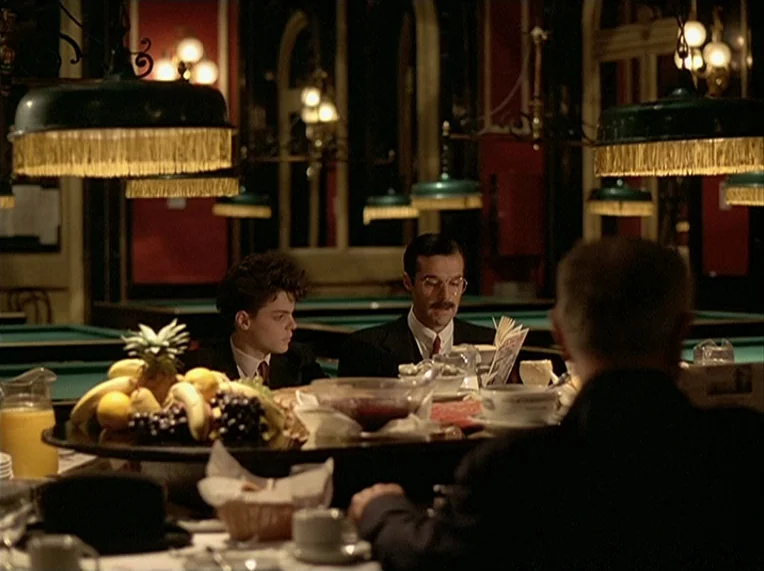 Detective (1985) movie still: medium — A dimly lit room features tables with dark green pool table surfaces and lamps. Two men in suits sit at a…