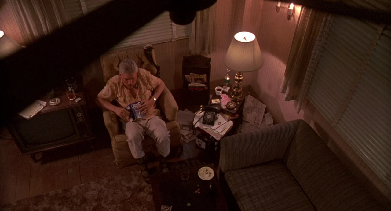 Night of the Creeps (1986) movie still: wide — A dimly lit room with vintage furniture and clutter scattered on various surfaces. An elderly man…