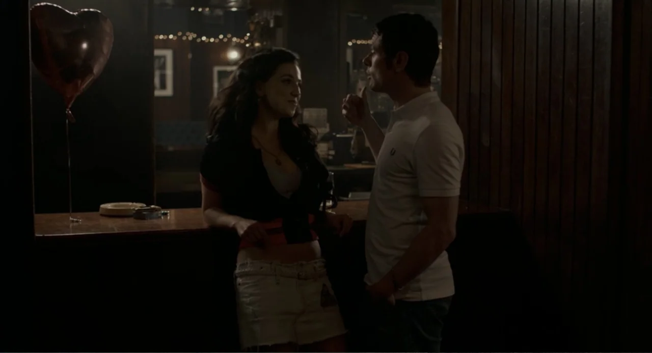 Back to Black (2024) movie still: medium — A dark dimly lit bar features a polished wooden service counter. A man and a woman stand facing each…