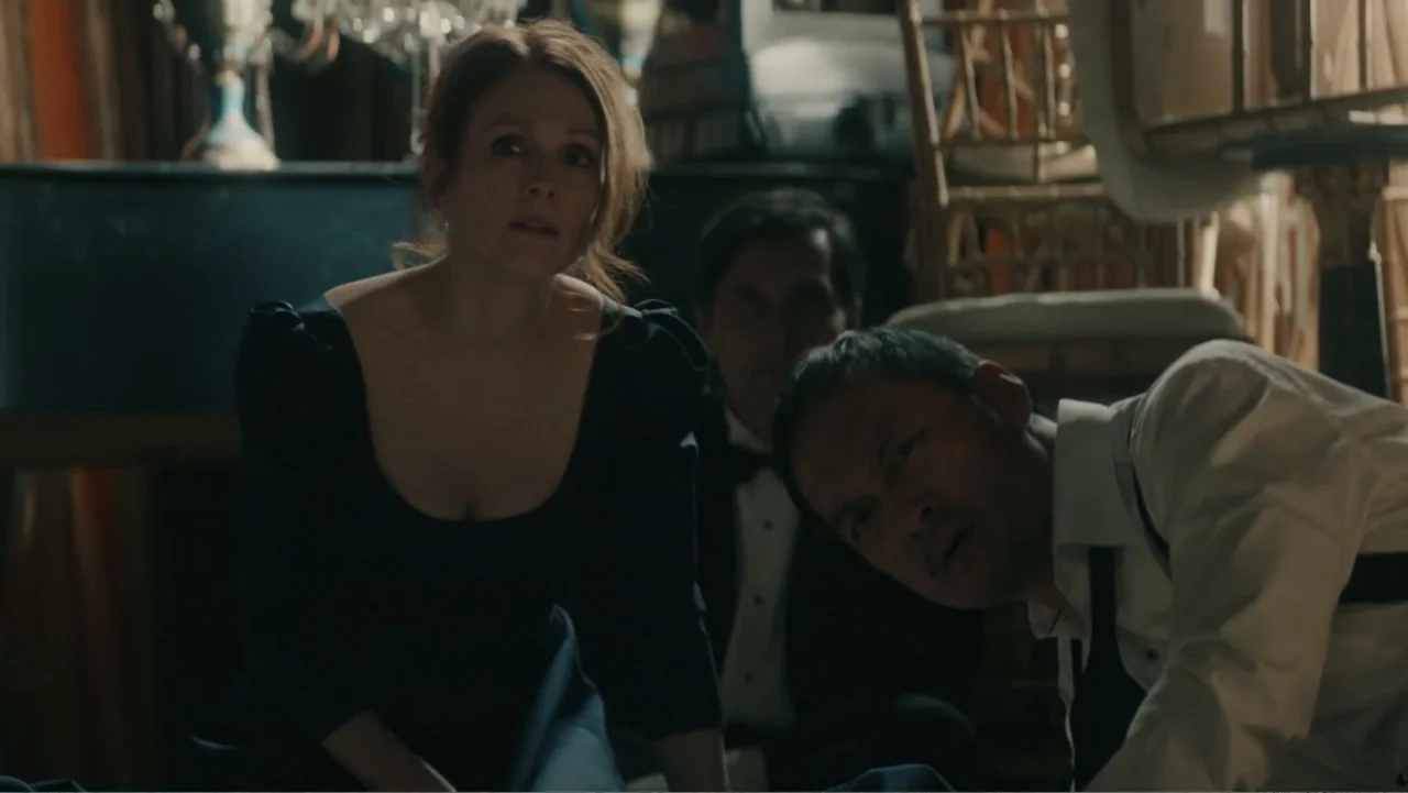 Bel Canto (2018) movie still: medium — A cluttered interior space contains stacked chairs and furniture under low ambient light. A woman and two men…