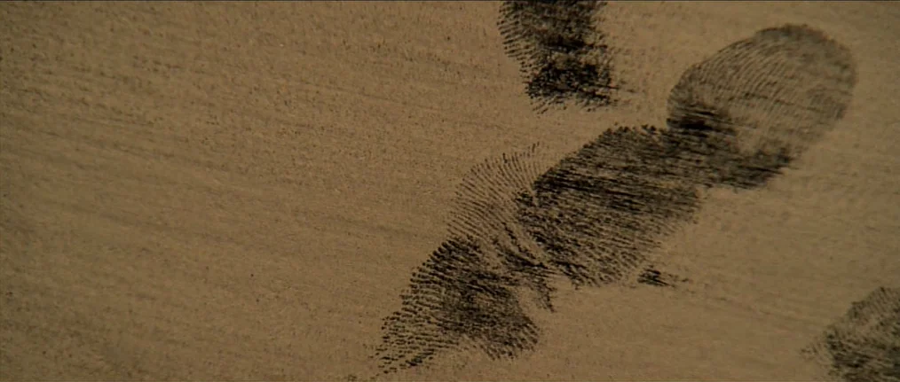 Se7en (1995) movie still: closeup — A close-up shot focuses on a textured, light brown surface. Several dark, distinct fingerprints are pressed into…