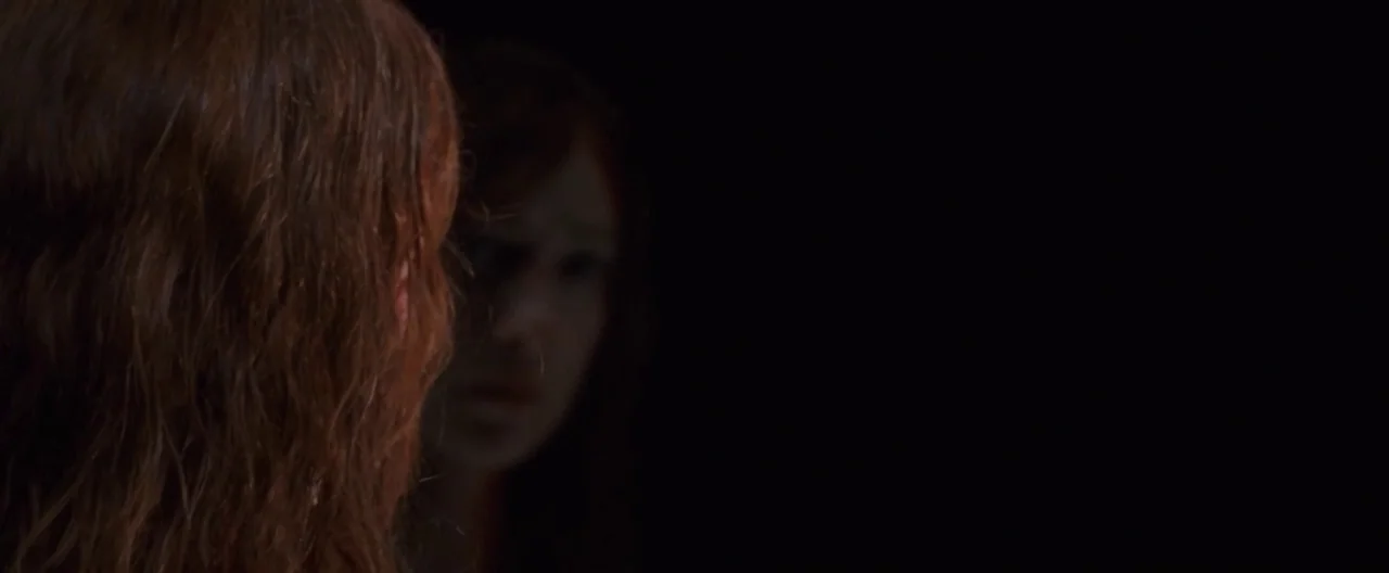 The Cabin In The Woods (2012) movie still: closeup — A woman stares into a dark reflective surface in a poorly lit room. Her face is partially…