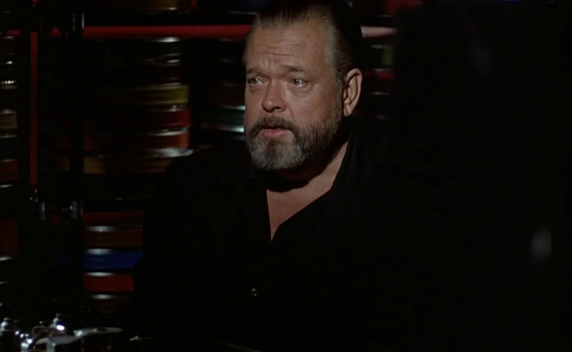 F for Fake (1973) movie still: medium — A dark space filled with stacked metal film reels in the shadows behind a man. An older man with a grey…