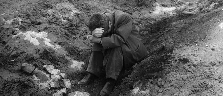 The Bad Sleep Well (1960) movie still: fullbody — A man sits inside a deep pit surrounded by uneven earth and rocky soil. The man in a formal suit…